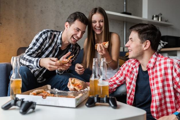 Happy young people eating pizza, drinking beer, having fun, friends party at home, hipster company together, two men one woman, smiling, positive, relaxed, hang out, laughing, 