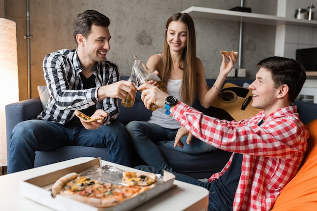 Happy young people eating pizza, drinking beer, having fun, friends party at home, hipster company together, two men one woman, smiling, positive, relaxed, hang out, laughing, 
