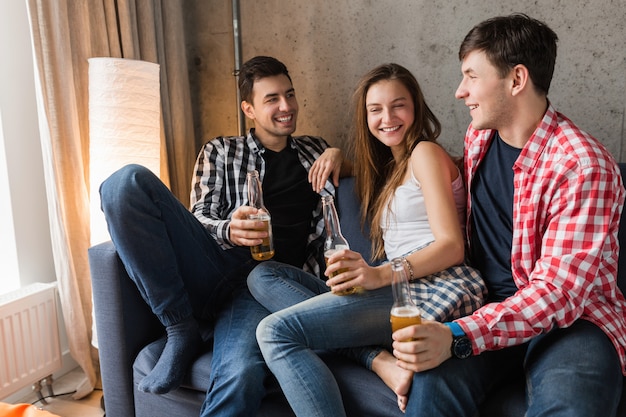 Happy young people sitting on sofa, drinking beer, close up hands toasting, having fun, friends home party, hipster company together, two men one woman, smiling, positive, relaxed, hang out, laughing
