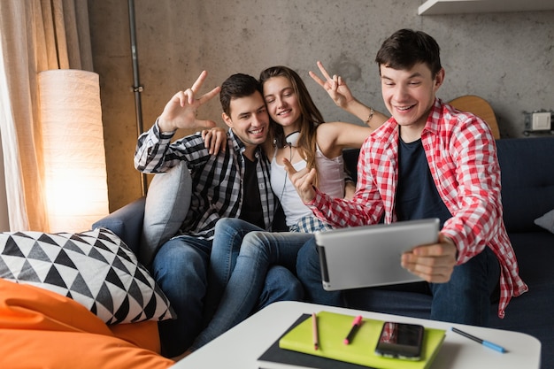 Happy young people using tablet, students having fun, making selfie photo, friends party at home, hipster company together, two men one woman, smiling, positive, online chat