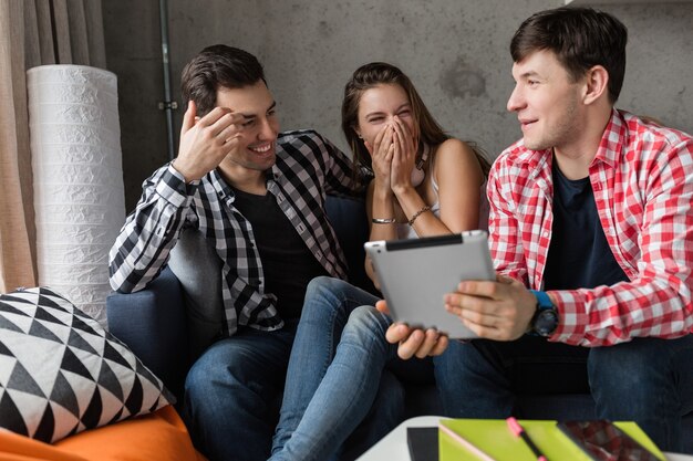 Happy young people using tablet, students learning, having fun, friends party at home, hipster company together, two men one woman, smiling, positive, online education