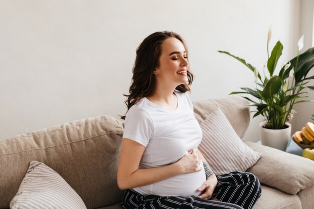 リビングルームで笑っている白いTシャツとパンツで幸せな若い妊婦