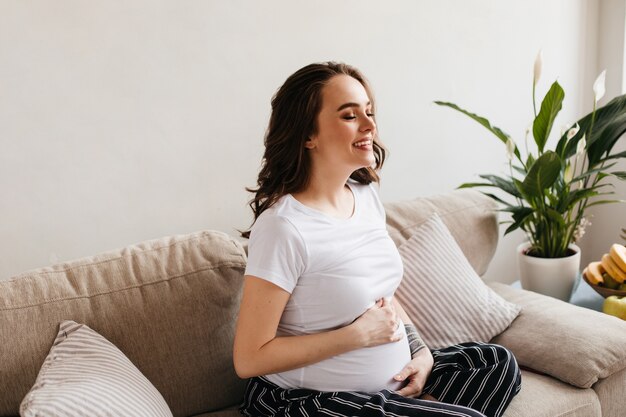 Счастливая молодая беременная женщина в белой футболке и штанах смеется в гостиной