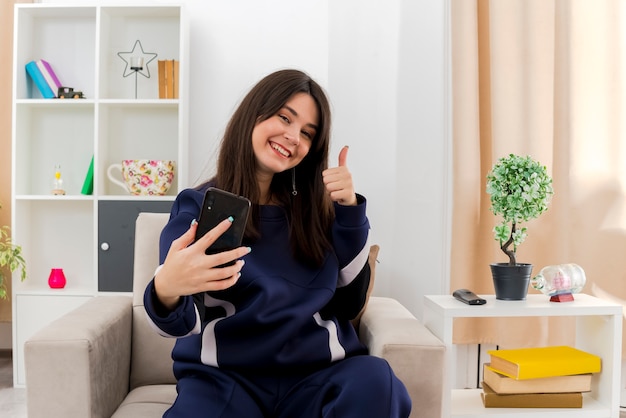 Счастливая молодая симпатичная кавказская девушка, сидящая на кресле в дизайнерской гостиной, протягивает мобильный телефон, показывая большой палец вверх и