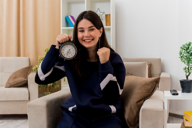 Happy young pretty caucasian woman sitting on armchair in designed living room holding alarm clock looking and doing yes gesture