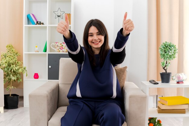 Happy young pretty caucasian woman sitting on armchair in designed living room showing thumbs up and looking
