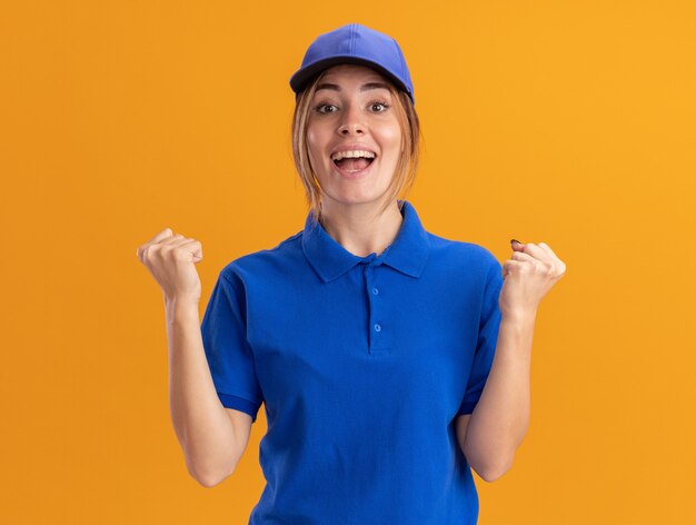 Happy young pretty delivery girl in uniform keeps fists on orange