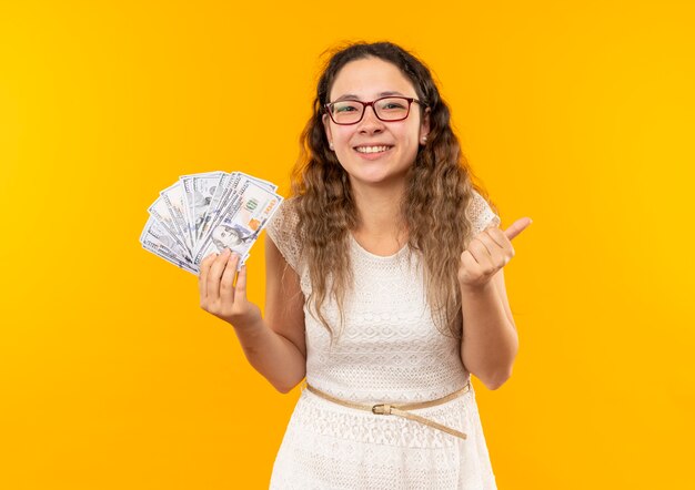 Happy young pretty schoolgirl wearing glasses holding money showing thumb up isolated on yellow  with copy space