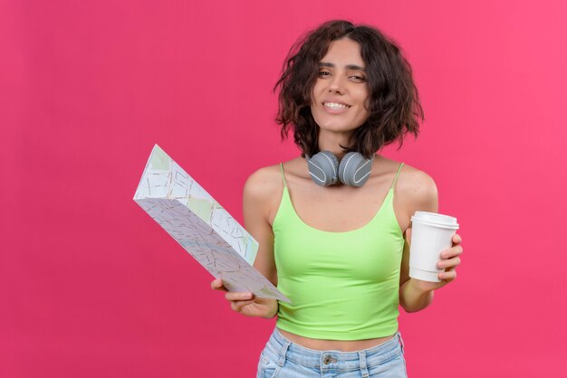 A happy young pretty woman with short hair in green crop top in headphones holding plastic cup of coffee and 