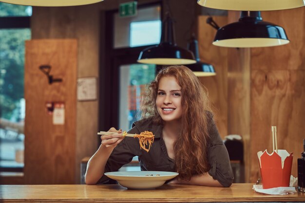 Happy young redhead female wearing casual clothes eating spicy noodles in an Asian restaurant.