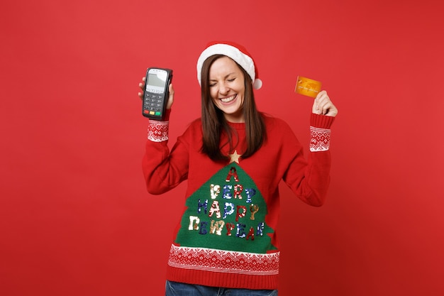Happy young santa girl hold wireless modern bank payment terminal to process and acquire credit card payments, card isolated on red background. happy new year 2019 celebration holiday party concept.