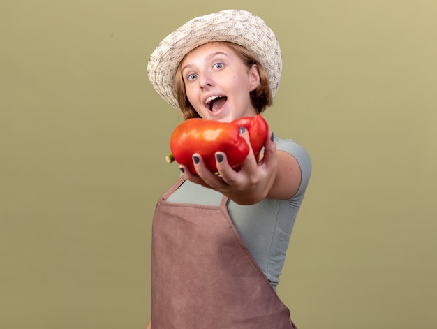 Happy young slavic female gardener wearing gardening hat holding red peppers