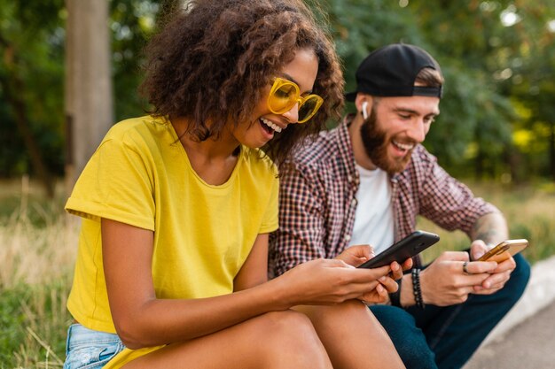 Happy young smiling friends sitting park using smartphones, man and woman having fun together