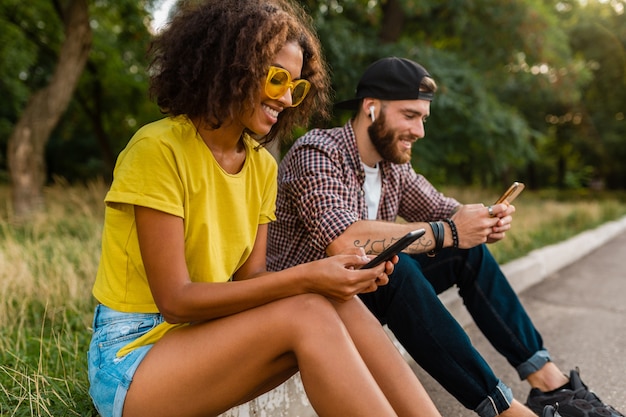 Счастливые молодые улыбающиеся друзья сидят в парке с помощью смартфонов, мужчина и женщина веселятся вместе