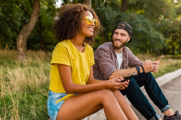 Happy young smiling friends sitting park using smartphones, man and woman having fun together