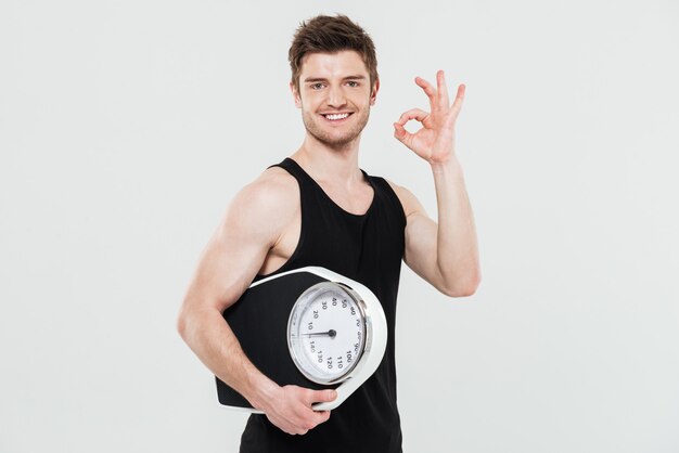 Happy young sportsman holding scales showing okay gesture
