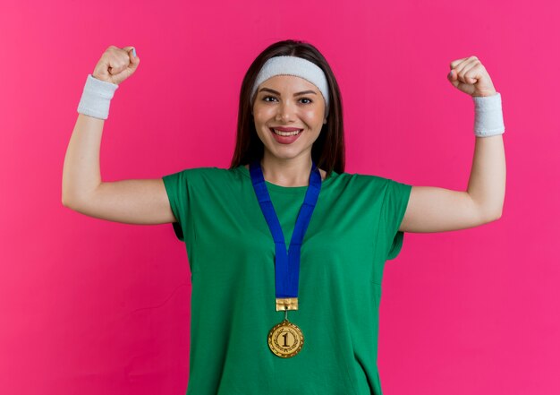 Счастливая молодая спортивная женщина, носящая повязку на голову и браслеты с медалью на шее, смотрит, делая сильный жест