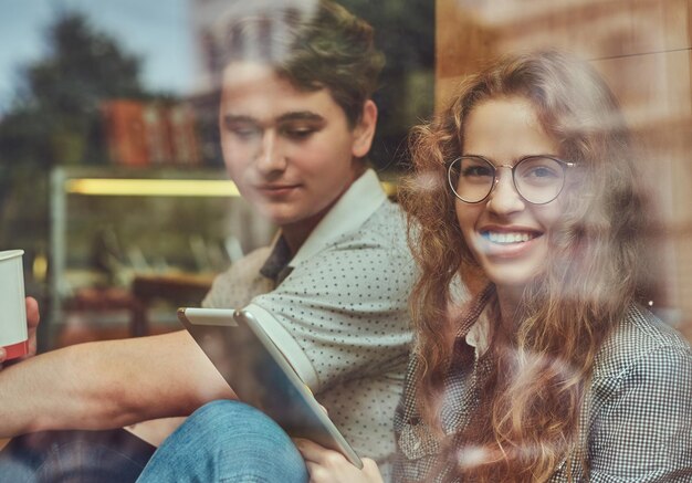 커피를 마시고 휴식 시간에 대학 캠퍼스의 창틀에 앉아 디지털 태블릿을 사용하는 행복한 젊은 학생들.