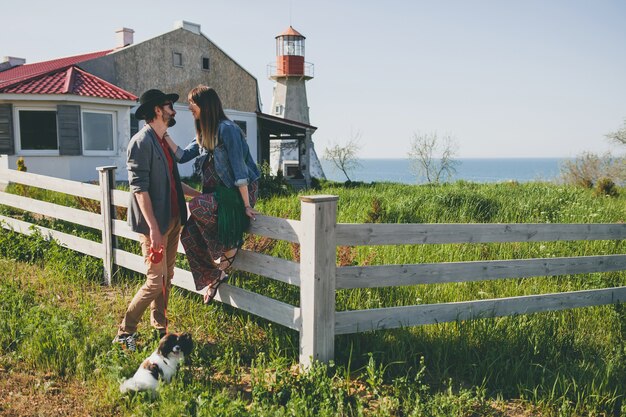 Happy young stylish hipster couple in love walking with dog in countryside, summer style boho fashion