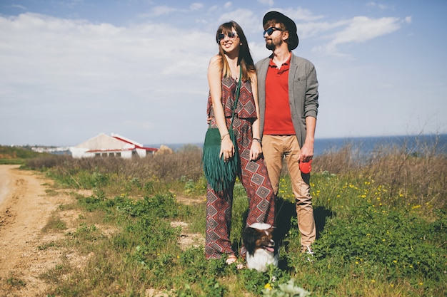 Happy young stylish hipster couple in love walking with dog in countryside