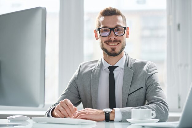 Happy young successful businessman in eyeglasses and suit sitting by desk in front of computer monitor