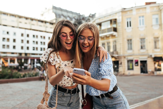 화려한 선글라스와 세련된 자른 블라우스를 입은 행복한 젊은 그을린 소녀들은 전화 화면을 보고 웃습니다. 금발과 갈색 머리 여성들은 메시지를 읽습니다