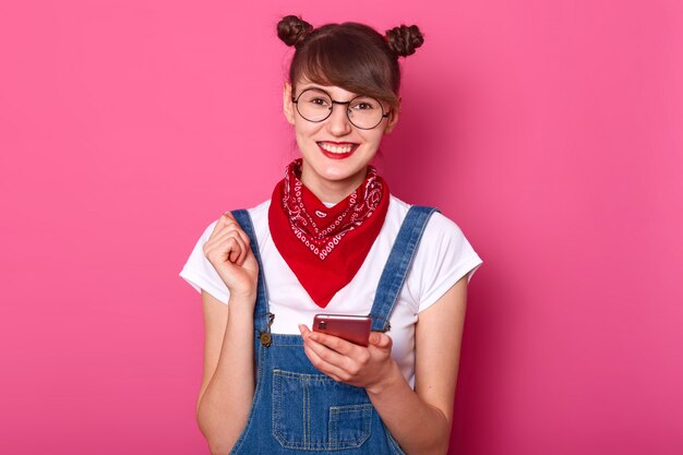 happy young teenager female with bunches, glasses, bandana on nack, dressed denim overalls and white t shirt, looks satisfied while holding mobile phone in hand isolated on pink.