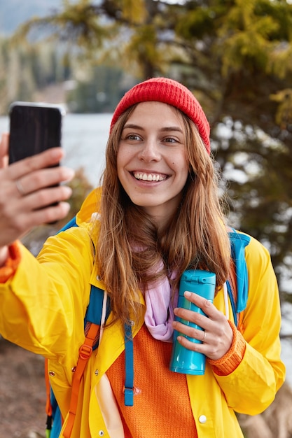 Happy young traveler smiles happily, does selfie with cell phone, dressed in yellow anorak, holds thermos with tea, has recreation in beautiful forest