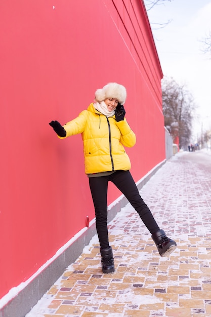 Happy young woman on a background of a red wall in warm clothes on a winter sunny day smiling and talking on the phone on a snowy city sidewalk