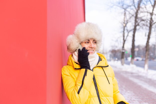 겨울 맑은 날에 따뜻한 옷을 입고 붉은 벽의 배경에 행복 한 젊은 여자 웃 고 눈 덮인 도시 보도에 전화로 이야기