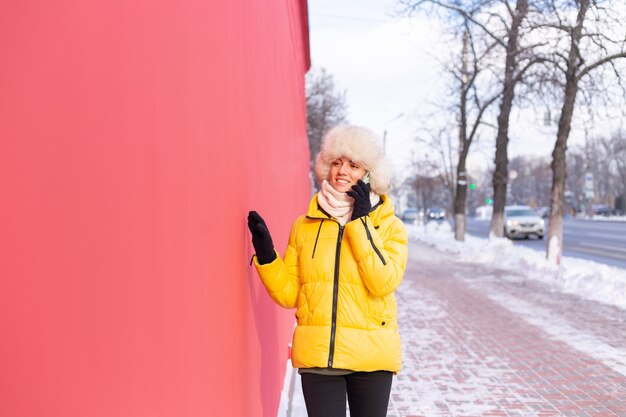Happy young woman on a background of a red wall in warm clothes on a winter sunny day smiling and talking on the phone on a snowy city sidewalk