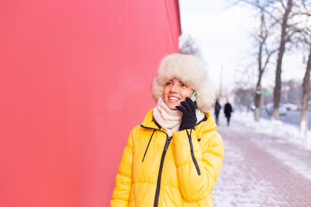 Happy young woman on a background of a red wall in warm clothes on a winter sunny day smiling and talking on the phone on a snowy city sidewalk