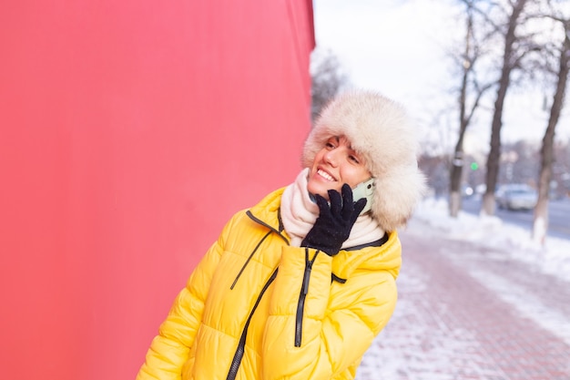 雪に覆われた街の歩道で笑顔と電話で話している冬の晴れた日に暖かい服を着て赤い壁の背景に幸せな若い女性