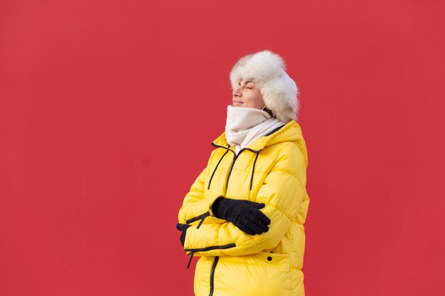 Happy young woman on the background of a red wall in warm clothes on a winter sunny day