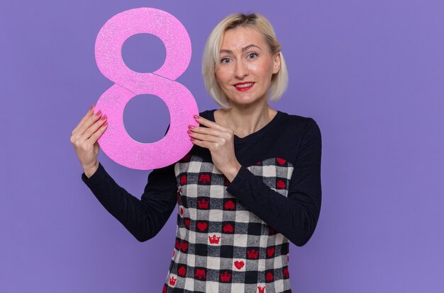 Happy young woman in beautiful dress holding number eight made from cardboard looking at front smiling cheerfully celebrating international women's day standing over purple wall