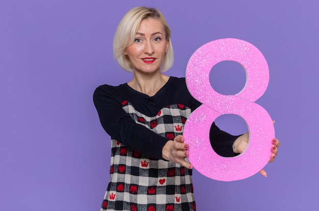 Free photo happy young woman in beautiful dress holding number eight made from cardboard looking at front smiling cheerfully celebrating international women's day standing over purple wall