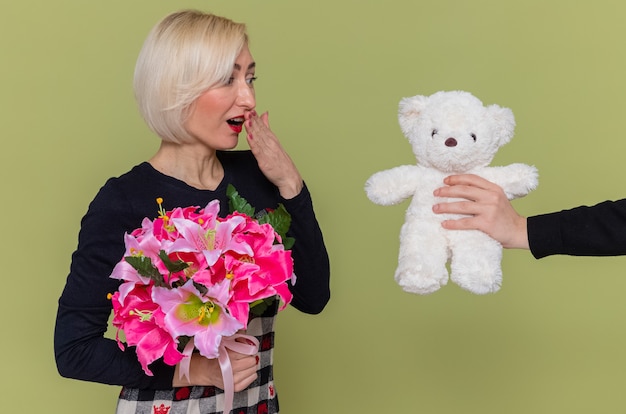 Happy young woman in beautiful dress with bouquet of flowers looking surprised smiling while receiving teddy bear as a gift celebrating international women's day standing over green wall