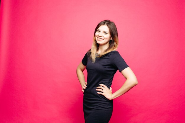 Happy young woman in black dress posing at studio and lauthing looking away Positivity emotionaly girl holding hand on waist standing against pink background Concept of shopping fashionbeauty