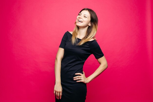 Happy young woman in black dress posing at studio and lauthing looking away Positivity emotionaly girl holding hand on waist standing against pink background Concept of shopping fashionbeauty
