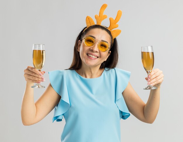 Happy young woman in blue top wearing funny rim with deer horns and yellow glasses holding two glasses of champagne smiling cheerfully 