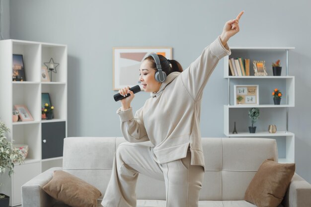 Счастливая молодая женщина в повседневной одежде с наушниками танцует на диване в домашнем интерьере, держа пульт, используя его как микрофон, поет весело