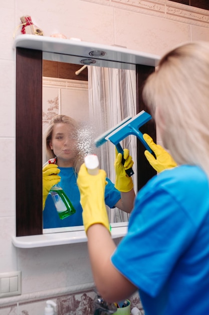 スプレーで掃除し、スキージを使用してバスルームで鏡を洗う幸せな若い女性家で働く黄色の保護手袋を着用したかわいいブロンドの女の子家事とハウスキーピングのコンセプト