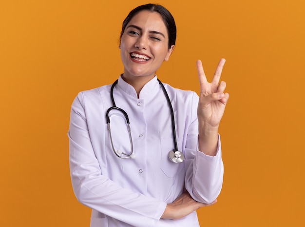 Felice giovane donna medico in cappotto medico con uno stetoscopio intorno al collo guardando la parte anteriore con un grande sorriso sul viso che mostra v-segno in piedi sopra la parete arancione