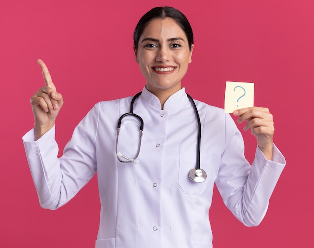 Happy young woman doctor in medical coat with stethoscope holding reminder paper with question mark pointing with index finger up smiling cheerfully standing over pink wall
