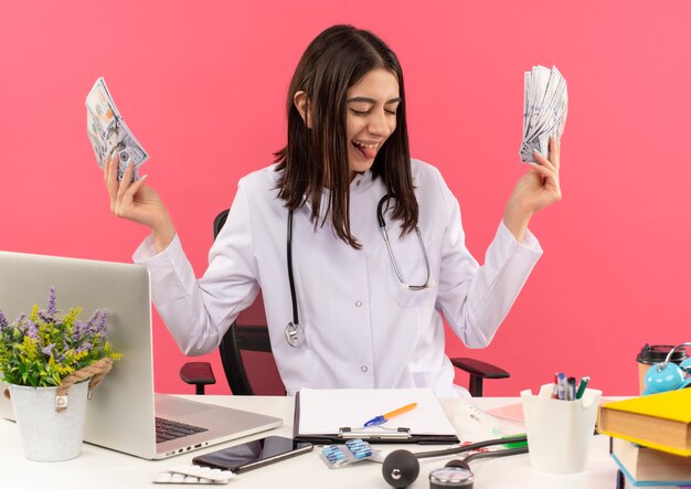 Счастливая молодая женщина-врач в белом халате со стетоскопом на шее держит деньги, весело улыбаясь, сидя за столом с ноутбуком над розовой стеной