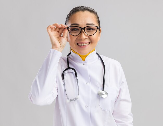 Happy young woman doctor in white medical coat wearing glasses with stethoscope around neck  with smile on face standing over white wall