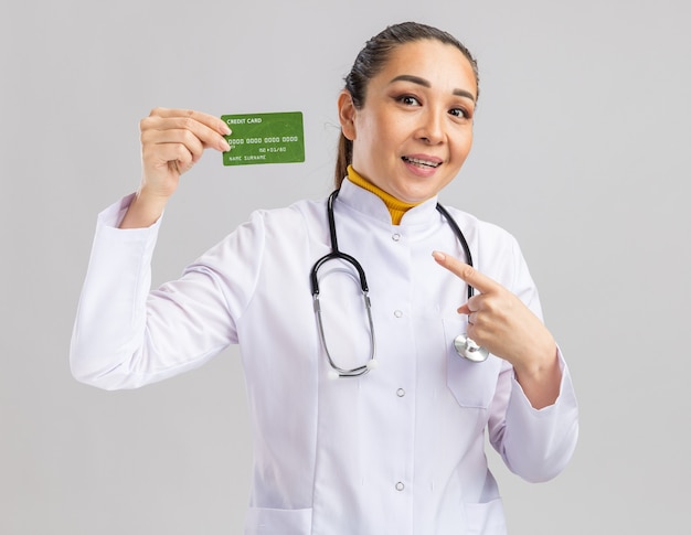 Happy young woman doctor in white medical coat with stethoscope around neck holding credit card pointing with index finger at it smiling cheerfully standing over white wall