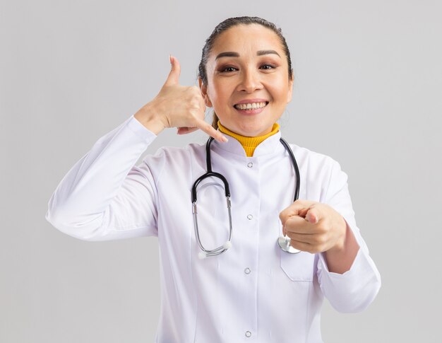 Happy young woman doctor in white medical coat with stethoscope around neck  smiling making call me gesture pointing with index finger at you standing over white wall