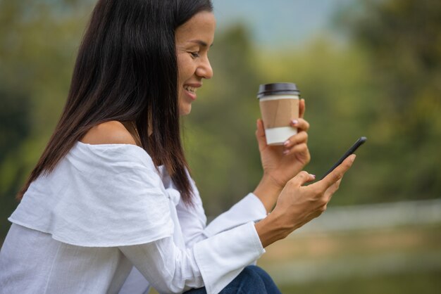 幸せな若い女がコーヒーを飲むと、スマートフォンを使用して