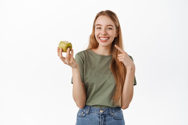 Giovane donna felice che mangia mela verde che indica il suo sorriso perfetto bianco che mostra i denti sani sbiancati che si levano in piedi contro il fondo bianco